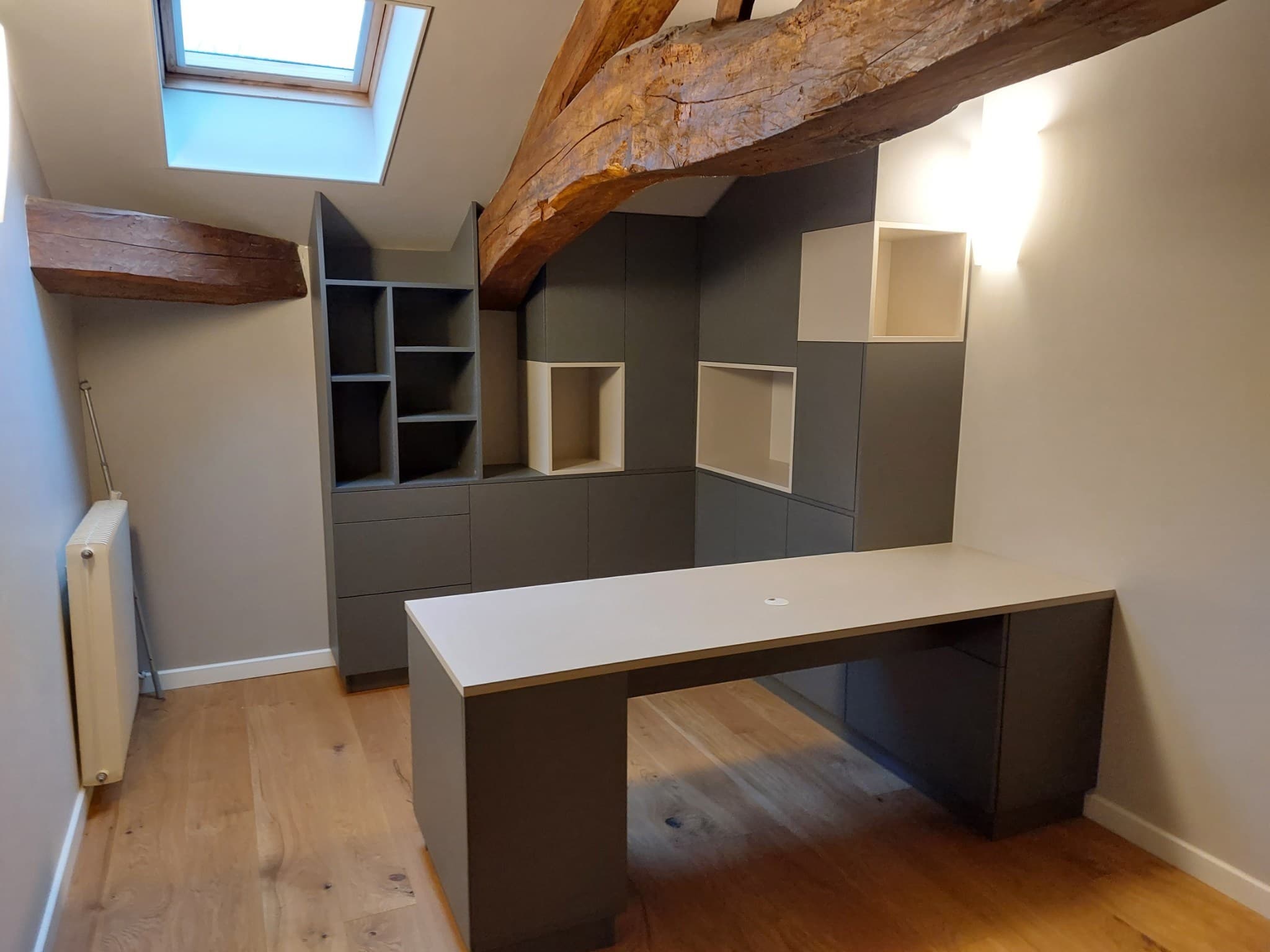Vue d’angle du bureau sous poutres apparentes, niches ouvertes et tiroirs anthracite, parquet chêne clair
