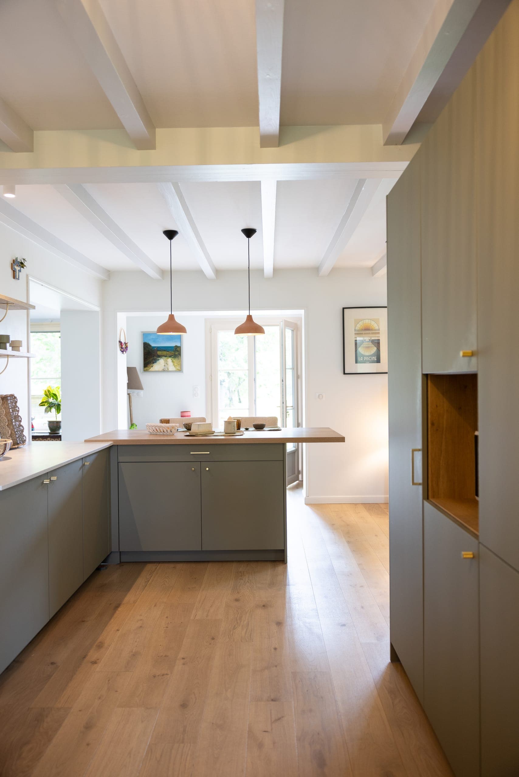 Perspective de la cuisine vers la pièce de vie, îlot gris-vert et hauts rangements toute hauteur